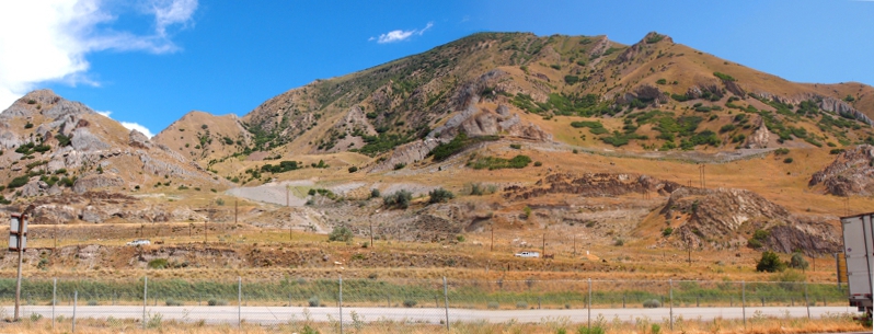 [Two photos stitched together showing grey rock, hillside with some greenery and some wheat-colored vegetation above the interstate.]